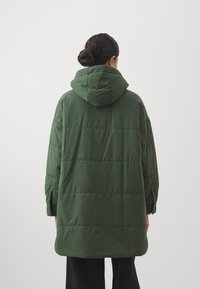 Grön puffjacka med huva, med ett kviltat mönster och långa ärmar. Tyget verkar slätt med en matt finish.