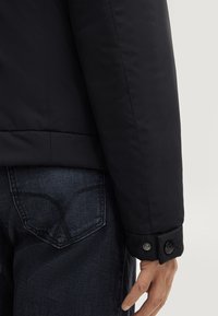 Giacca nera imbottita con una texture liscia, dotata di polsini con bottone e design minimalista, abbinata a jeans in denim scuro.