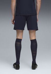 Pantalones cortos deportivos de color marino y calcetas hasta la rodilla con patrones a rayas. Zapatillas de fútbol negras con un logo en el lateral. Tela con textura visible.
