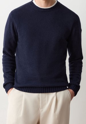 Mężczyzna ubrany w ciemnogranatowy sweter z okrągłym dekoltem, na białej podkoszulce, w jasnobeżowych spodniach, z częściowo wsuniętymi rękami do kieszeni, na jednolitym tle.