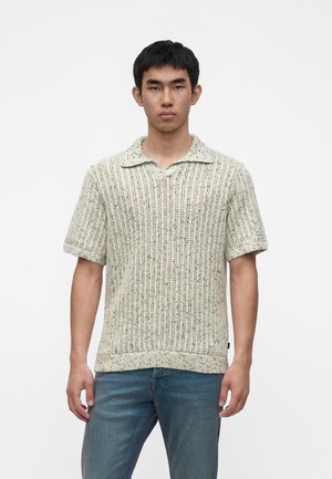Jeune homme portant un pull en maille à manches courtes avec un col lâche, associé à un jean bleu, debout devant un fond blanc uni.