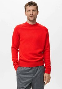 Rdeča pletena majica s krožnim izrezom in rebrastimi manšetami. Ima nekoliko ohlapen kroj in mehak material, kombinirana s sivo hlačami.