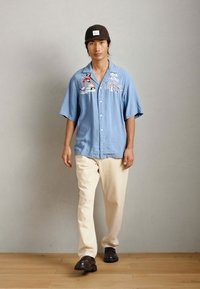 Ανοιχτό μπλε κοντομάνικο πουκάμισο με λουλουδάτη κεντημένη διακόσμηση, κρέμα παντελόνια και σκούρα παπούτσια. Το πουκάμισο διαθέτει χαλαρή γραμμή και κουμπί κλεισίματος.