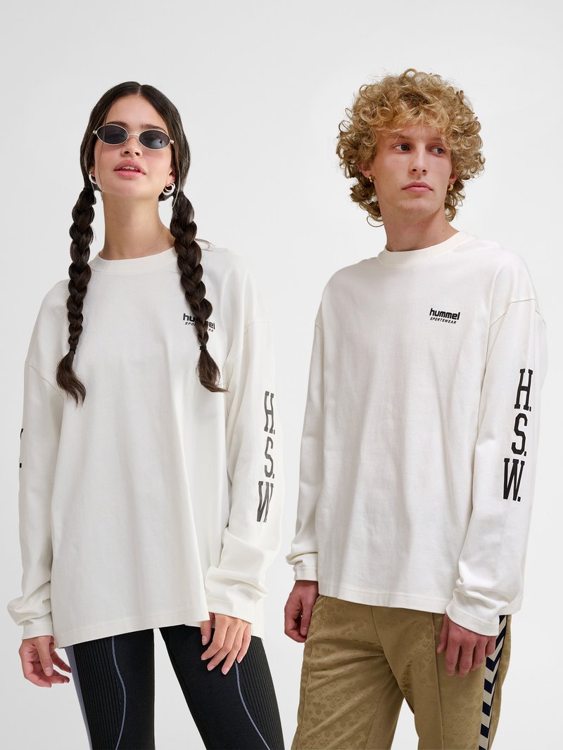 Dos adultos jóvenes llevan camisetas blancas de manga larga con "H.S.W." en las mangas; uno tiene el pelo trenzado y gafas de sol, el otro tiene el pelo rizado.