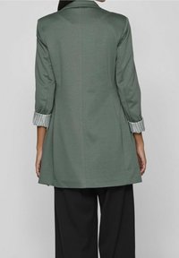 Zelené sako s dlouhými rukávy a proužkovanými manžetami. Na míru střižené s hladkou texturou, vhodné k širokým černým kalhotám.