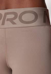 Detailní záběr osoby v těsných legínách v barvě taupe s širokým pasem, na kterém jsou velká tmavě taupe písmena.