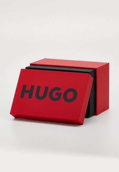 Boîte rouge avec une couche intérieure noire, arborant un texte noir en gras "HUGO" sur le devant. Design géométrique simple, texture lisse.