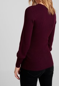 Burgunderfarbener Rippstrickpullover mit langen Ärmeln, figurbetontem Design und rundem Halsausschnitt, kombiniert mit schwarzen Jeans. Glatte Textur, ohne Muster.