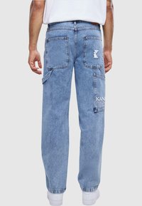 Jeans de mezclilla azul claro con corte relajado, que cuentan con dos bolsillos traseros y un bolsillo de carga con detalle de logo. Costuras blancas y dobladillos simples.