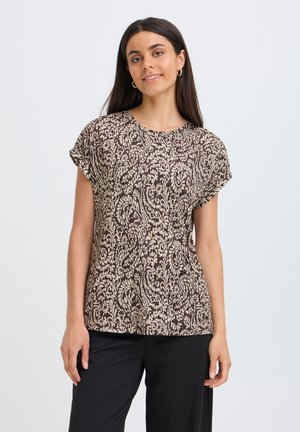 Joven mujer con cabello largo y oscuro, vestida con una blusa de manga corta marrón y beige con estampado y pantalones negros, de pie contra un fondo liso.