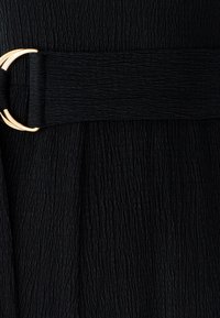 Sort tekstureret stofbeklædning med et bredt bælte med en guldfarvet rund spænde. Overfladen har et krøllet mønster.