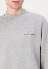 Homme portant un sweat-shirt gris clair à col rond avec un petit texte noir « SAMSØE SAMSØE » sur la poitrine gauche.