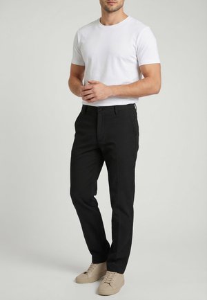 Hombre con camiseta blanca de manga corta, pantalones negros y zapatillas beige, de pie con las manos entrelazadas al frente.