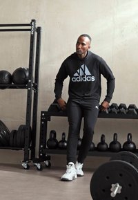 Sudadera negra de Adidas con logotipo gris, pantalones ajustados negros y zapatillas deportivas blancas; pesas y kettlebells en el fondo.