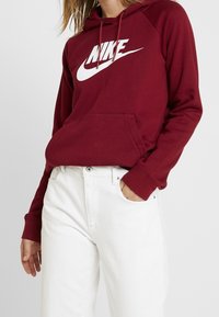 Kvinna iklädd en vinröd Nike-hoodie med vitt logotyp och vita högmidjade jeans, en hand i fickan, mot en ljus bakgrund.