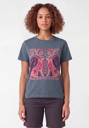 Vrouw met krullend haar die een grijs T-shirt draagt met een symmetrische roze en rode afbeelding van twee gestileerde honden die elkaar aankijken.