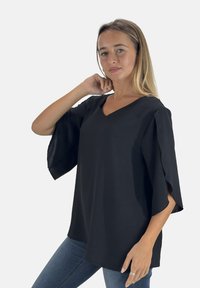 Zwarte top met korte mouwen, V-hals en uitlopende mouwen. De stof lijkt glad met een lichte glans. Gecombineerd met een blauwe spijkerbroek.