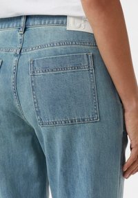 Jasnoniebieskie dżinsy z denimu o średnim stanie, z dwiema tylnymi kieszeniami i białą naszywką z logo na pasku. Miękka tekstura i prosty krój.