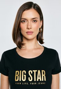 Černé tričko s krátkými rukávy a kulatým výstřihem, na kterém je zlatý potisk s nápisem „BIG STAR TVŮJ ŽIVOT. TVÉ DŽÍNY.“