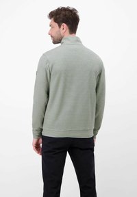 Zielony prążkowany sweter z wysokim kołnierzem, długimi rękawami i dopasowanym dolnym brzegiem. Tył ma subtelną fakturę oraz naszywkę na rękawie.