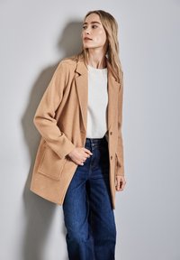Abrigo de mezcla de lana color camel con un diseño entallado, que presenta solapas con muesca, dos bolsillos frontales y un ajuste relajado, combinado con jeans de mezclilla.