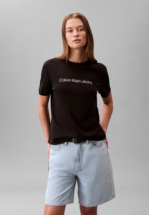 T-shirt noir en coton avec le texte blanc "Calvin Klein Jeans", assorti à un short en denim bleu clair. Le mannequin pose les mains dans les poches.