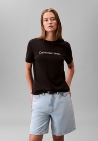 Czarna bawełniana koszulka z białym napisem "Calvin Klein Jeans", zestawiona z jasnoniebieskimi szortami dżinsowymi. Model stoi z rękami w kieszeniach.