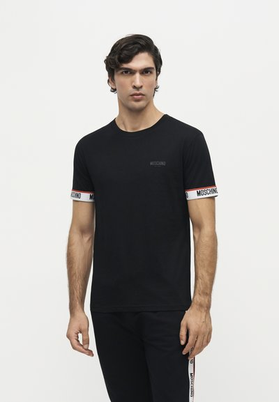Homme portant un t-shirt Moschino noir avec des bordures blanches, rouges et noires sur les poignets des manches, debout devant un fond blanc uni.