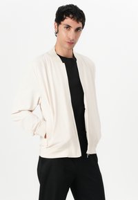 Light beige bomber jacket with a smooth texture, ribbed cuffs, and a front zipper. Worn over a black shirt, set against a white background.