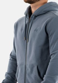 Lichte blauwe zip-up hoodie, gemaakt van zachte stof. Heeft een voorzak, verstelbare trekkoorden en een logo op de borst.