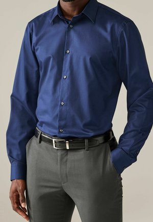 Homme portant une chemise ajustée bleu foncé à manches longues boutonnée, pantalon gris et ceinture noire, une main dans la poche.