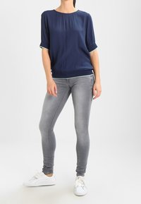 Námořnická modrá halenka s krátkým rukávem, kulatým výstřihem a ohrnutými manžetami, kombinovaná s šedými skinny džíny a bílými teniskami. Hladká textura, neformální design.