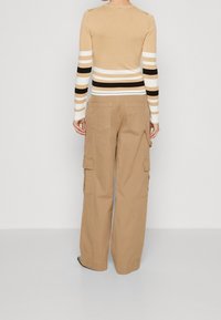Haut beige à manches longues avec des rayures blanches, noires et beiges ; associé à un pantalon cargo. Le pantalon est kaki, avec des poches et un fit ample.