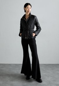 Black quilted jacket with a high collar, drawstring waist, and gold-tone hardware, paired with flared black pants and black footwear.