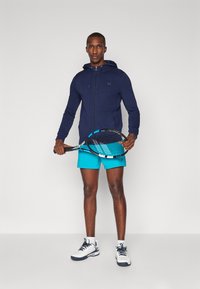 Mornarska zip-up dukserica s vezicama, uparena s jarkoplavim šortsem. Drži crni teniski reketa s plavim naglascima. Bijele sportske tenisice.