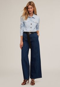 Camicia azzurra chiara con due tasche sul petto, abbinata a jeans a vita alta di colore blu scuro e cintura nera, accentuata da sandali con tacco.