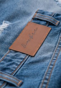 Brunt lædermærke med kursiv tekst på blå cowboybukser i denim, der viser syning og et lille slidt område.