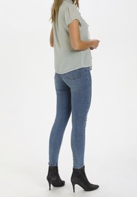 Blouse verte clair à manches courtes avec des accents de volants, associée à un jean slim bleu et des bottines noires, vue de dos.