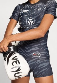 Maglia UFC a maniche corte in blu scuro con un motivo geometrico simile a un camuffamento. Il design include un logo e il marchio, abbinati a guantoni da boxe bianchi.