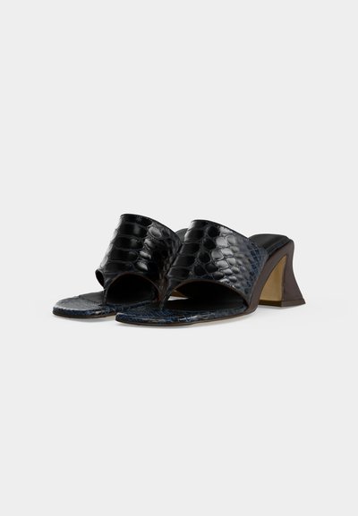 Μαύρα ανοιχτά σανδάλια mule με υφή κροκοδείλου και φαρδιές τακούνια σε απλό ανοιχτό φόντο.