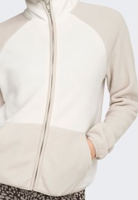 Veste zippée en polaire blanc et beige clair, avec manches raglan, poches latérales et texture douce. Détail de fermeture éclair devant.