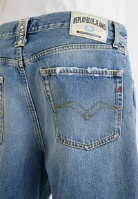 Gai zila džinsu auduma bikses ar nodilušu tekstūru, ar uzlīmi "REPLAY BLUE JEANS" un divām aizmugures kabatām ar šuvju detaļām.