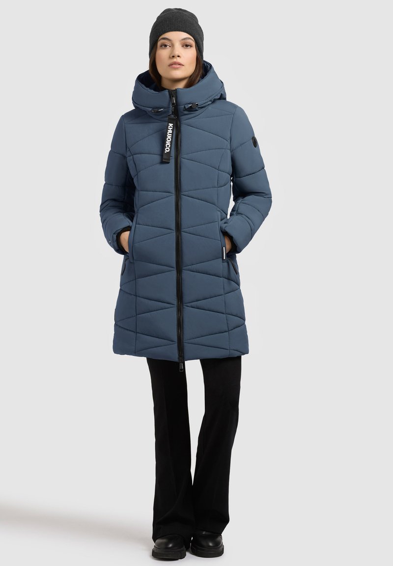 Modrá izolovaná bunda s geometrickým prešívaným dizajnom, vysokým golierom, predným zipsom a bočnými vreckami. Spárovaná s čiernymi nohavicami a čiapkou.