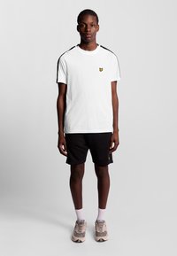 Vit kortärmad t-shirt med svarta axelremmar och gul logotyp, tillsammans med svarta shorts och beige sneakers. Modellen står mot en enfärgad bakgrund.