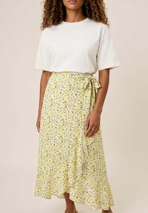 Mujer que lleva una blusa blanca lisa de manga corta y una falda cruzada amarilla con estampado floral y bajo con volantes atada a la cintura.