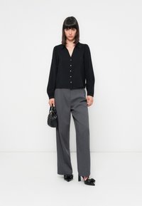 Blusa preta de mangas longas com decote em V e botões dourados, combinada com calças cinzentas de perna larga e sapatos de salto pretos. Segura uma pequena mala preta.