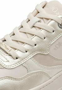 Beige sneaker featuring a mix of smooth leather and textured fabric, with metallic accents and flat laces, and a stitched design pattern.