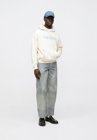 Beige Kapuzenpullover mit dem Schriftzug "LACONTE", hellblaue Mütze, weite graue Jeans und schwarze Schuhe. Der Kapuzenpullover verfügt über eine Kängurutasche und Kordeln.