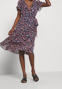 Kobieta w granatowej sukience w kwiaty z wiązaniem i czarnych klapkach bez pięty stoi na tle jednolitego jasnego tła.