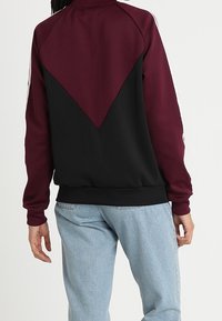 Vinröd och svart jacka med en färgblockdesign, med en triangulär baksektion, ribbade ärmslut och synliga sömnadsdetaljer.